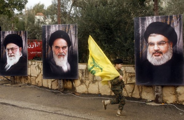 A boy carried a Hezbollah flag in Jibchit, Lebanon, in February as he ran past portraits of, from left, Ayatollah Ali Khamenei, the current supreme leader of Iran; Ayatollah Ruhollah Khomenei, the leader of Iran's Islamic Revolution; and Hassan Nasrallah, the leader of Hezbollah. Credit Mahmoud Zayyat/Agence France-Presse — Getty Images